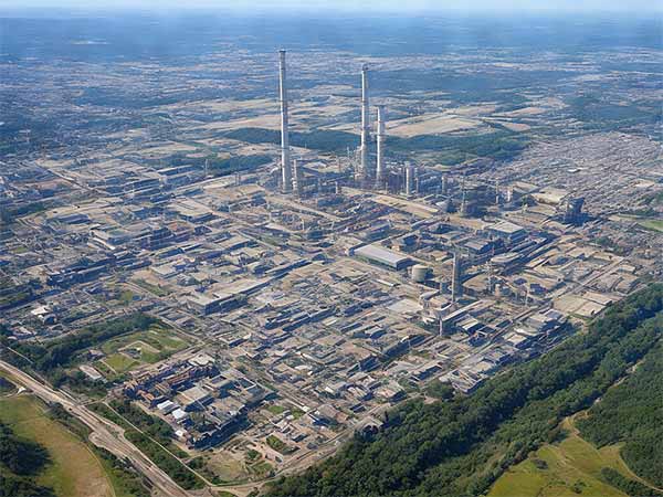 Aerial view of a large industrial complex with several buildings and towering chimneys, surrounded by green fields.