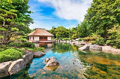 Tranquil Japanese garden with a thatched-roof gazebo by a clear pond, surrounded by lush greenery and rocks, under a bright blue sky with scattered clouds.