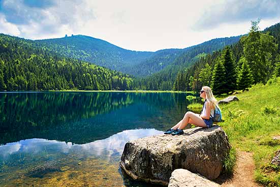 A person with a cap and backpack sits on a large rock next to a quiet lake, surrounded by wooded hills (Bavarian Forest National Park).