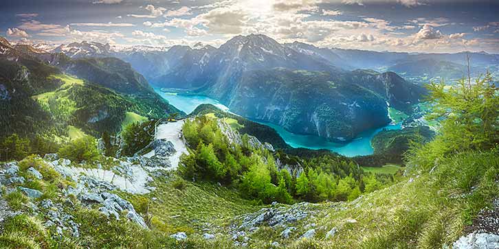 A panoramic view of a mountain landscape with lush greenery, rocky peaks and a turquoise lake under a partly cloudy sky.