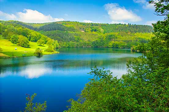 A tranquil lake surrounded by lush green hills and trees under a clear blue sky with a few scattered clouds.