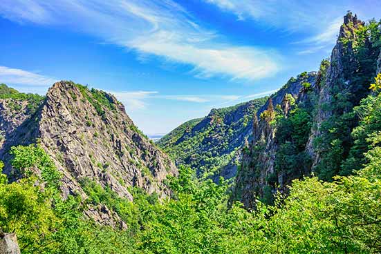 A vibrant landscape with a green valley flanked by steep, rocky mountains under a bright blue sky with thin clouds.