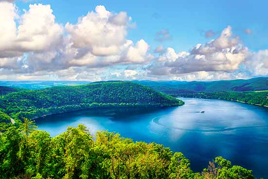 A picturesque view of a tranquil lake surrounded by lush green hills under a blue sky with scattered clouds. A boat can be seen on the lake.