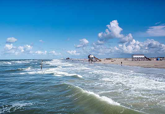 A sandy beach with gentle waves, blue skies and white clouds. Along the coast, buildings and people can be seen, including a building on stilts.