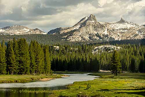 Unicorn Peak With Summer Snow Stands High Over Tuolomne River A tranquil landscape with a river meandering through a green forest, with mountains in the background under a cloudy sky.