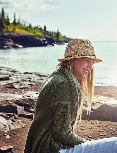test Inner peace through connection with nature: Woman in a green sweater and straw hat sits smiling on the rocky shore, water and trees can be seen in the background.