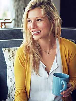 test A person with long hair sits on a sofa, holds a blue cup of coffee, wears a yellow cardigan and a white shirt, smiles and looks to the side.
