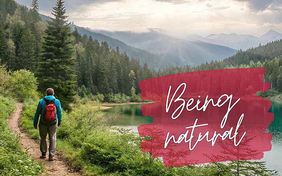 A person wearing a red backpack walks along a forest path by a lake. In the background, majestic mountains rise, while "Being natural" is written in large letters.